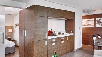 Ein modernes Hotelzimmer mit einem Holzkabinett, einer kleinen Küchenzeile, einer Essecke mit Tisch und Stühlen sowie einem Bett mit Nachtschrank.