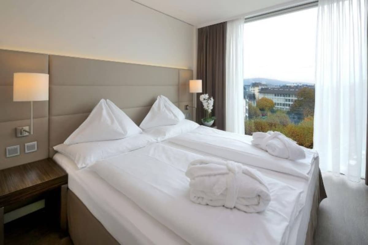 Habitación de hotel con una cama grande, dos mesitas de noche, una lámpara, un jarrón de flores y vistas a la ciudad.