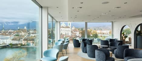 Un área de descanso moderna con asientos y un amplio ventanal con vistas a la ciudad y al río.