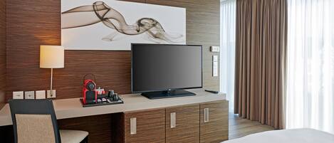 A hotel room with a bed, a TV on a wooden cabinet, a desk with a chair, and a large abstract painting on the wall.
