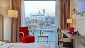 A hotel room with a bed, a red armchair, a dining table with a coffee maker, and a view of a historic building across a body of water. - H4 Hotel Solothurn (Solothurn)