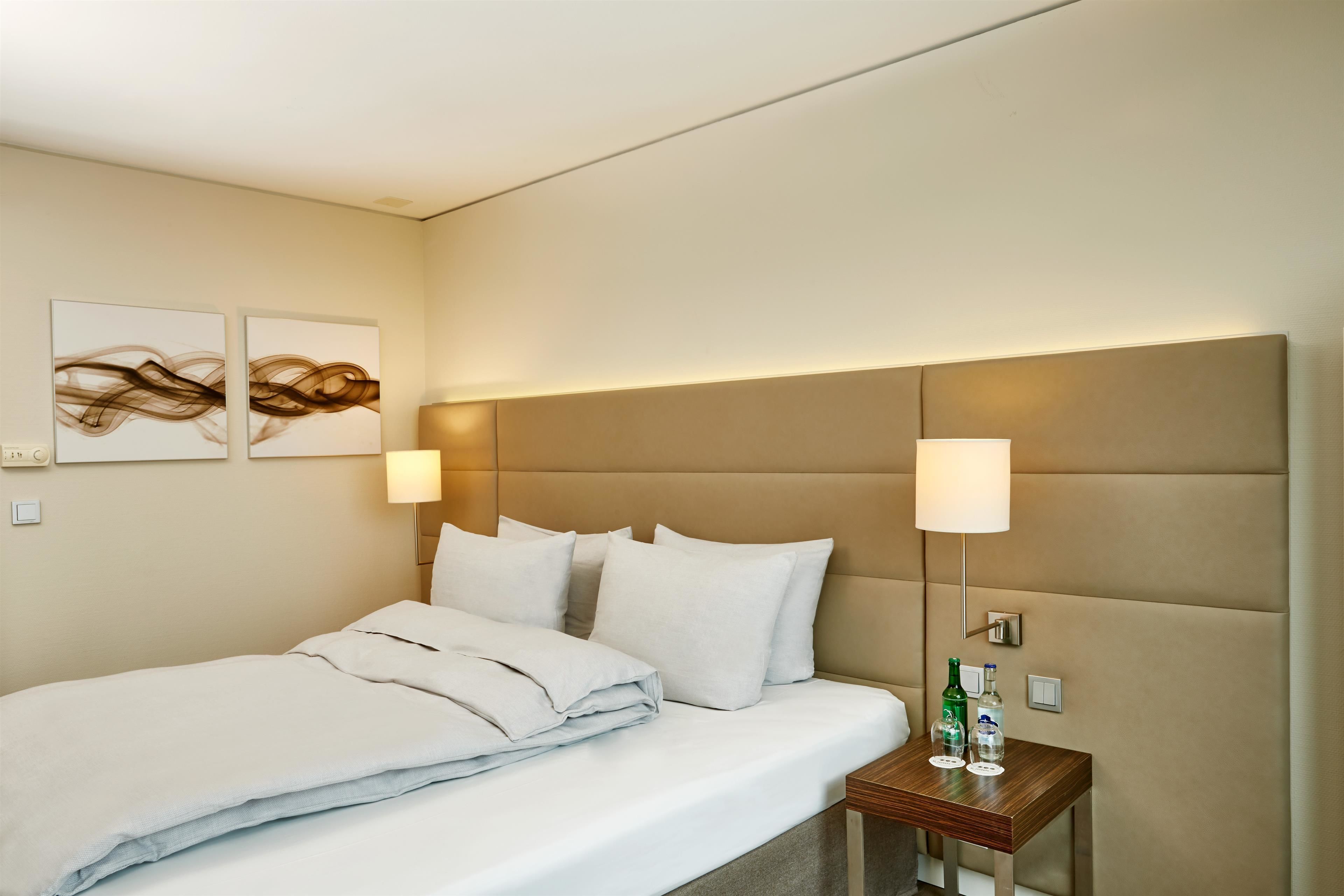 A hotel room with a bed, two wall-mounted lamps, a small wooden nightstand, and two abstract wall art pieces.
