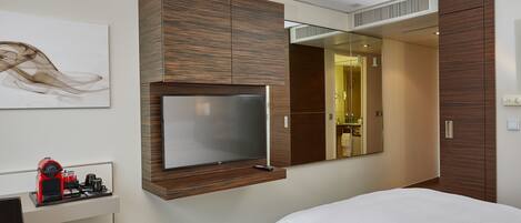 Une chambre d’hôtel moderne équipée d’un lit, d’un téléviseur fixé sur un panneau en bois, d’une machine à café et d’un miroir qui reflète la salle de bain.