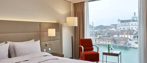 A hotel room with a large bed, a red armchair, a small table with a bottle, and a view of a cityscape and water.
