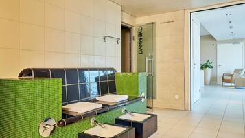 Um banheiro moderno com parede revestida em azulejos verdes, box de vidro e área de estar com um banco e duas pias.