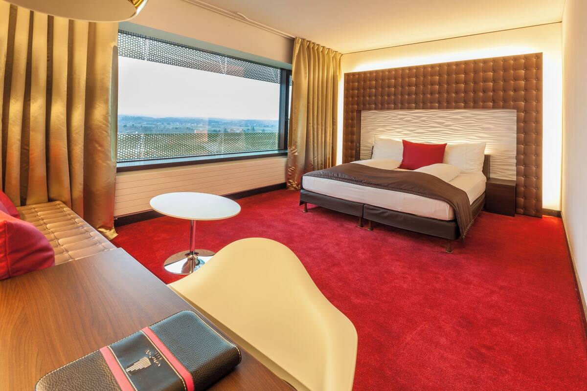 A hotel room with a large bed, a small round table, a sofa, and a view of the city through a large window.