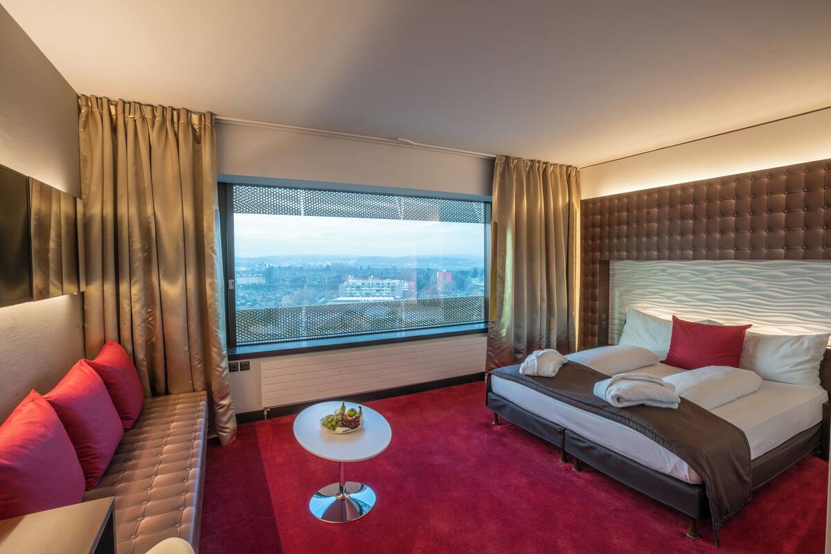 A hotel room with a large window, a bed with a headboard, a sofa, a small round table, and a red carpet.