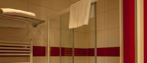 Ducha con cerramiento de vidrio, toalla blanca en la puerta, un perchero blanco y una franja roja en la pared.
