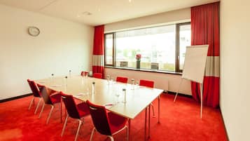 A conference room with a long table, red chairs, a whiteboard, and a window with red curtains.