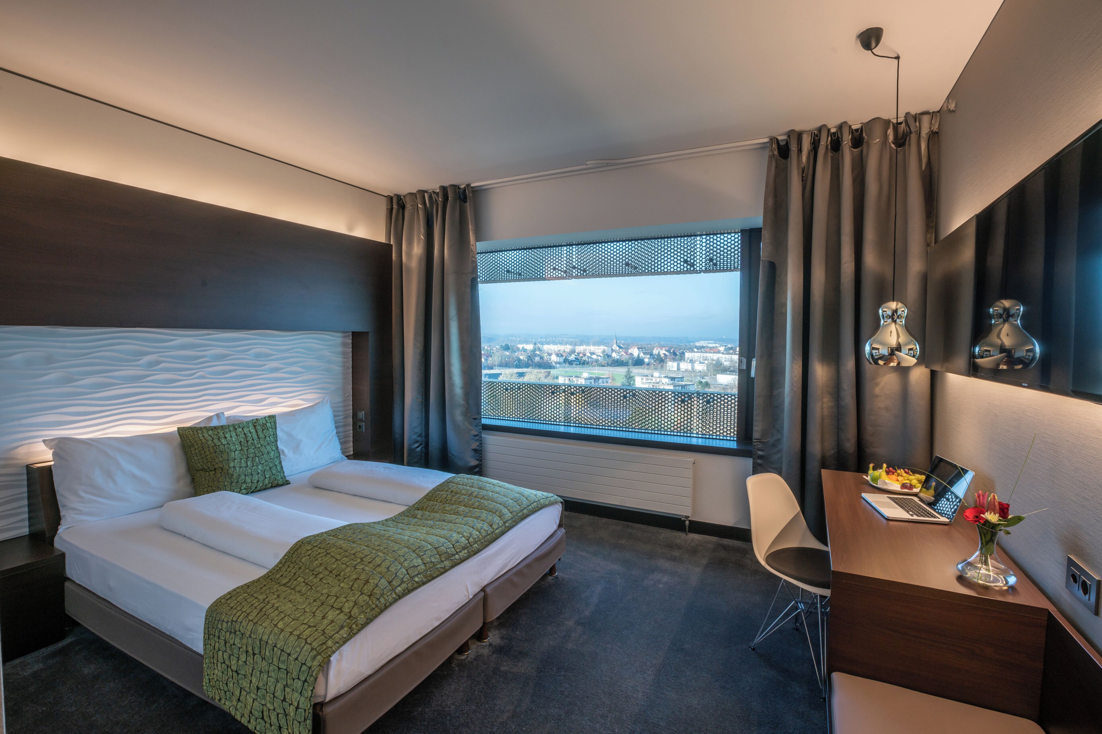 A hotel room with a large bed, a desk with a laptop, and a view of a cityscape through a window.