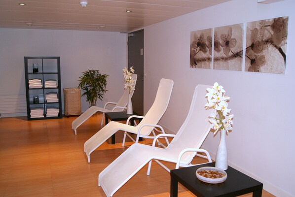 A room with three white lounge chairs, a black table with a bowl of snacks, a white vase with flowers, a shelf with folded towels, and three flower paintings on the wall.