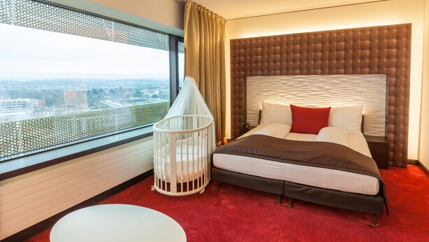 A hotel room with a large bed, a crib, a round table, and a city view through a large window.