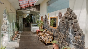 Un pasillo con suelo de piedra, una puerta de vidrio que da a una habitación, un banco de madera con cojines y plantas en macetas.