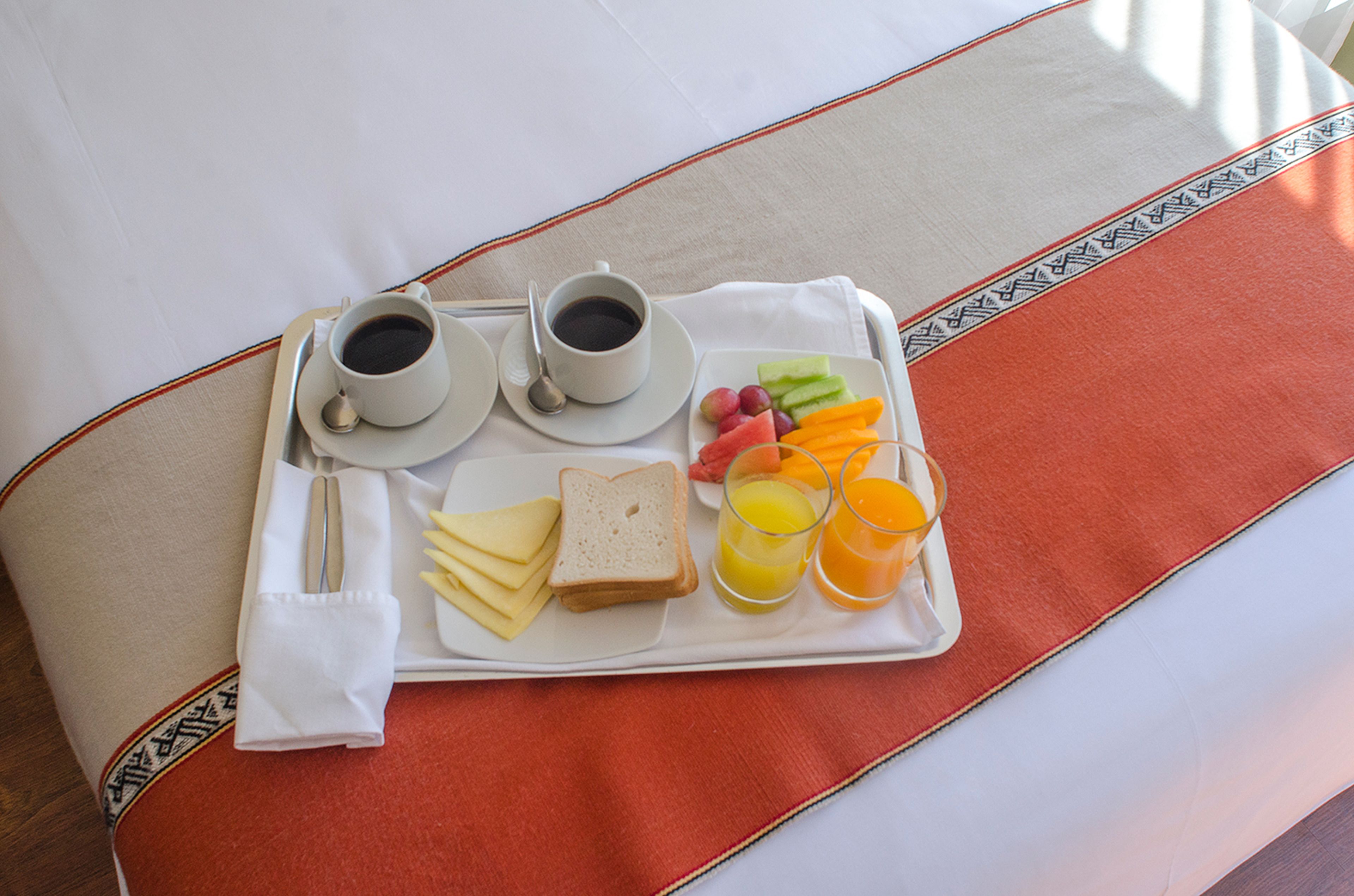 Ein Frühstücksbrett mit zwei Tassen Kaffee, Käse, Brot, Obst und Orangensaft auf einem Bett mit einem gemusterten Überwurf.