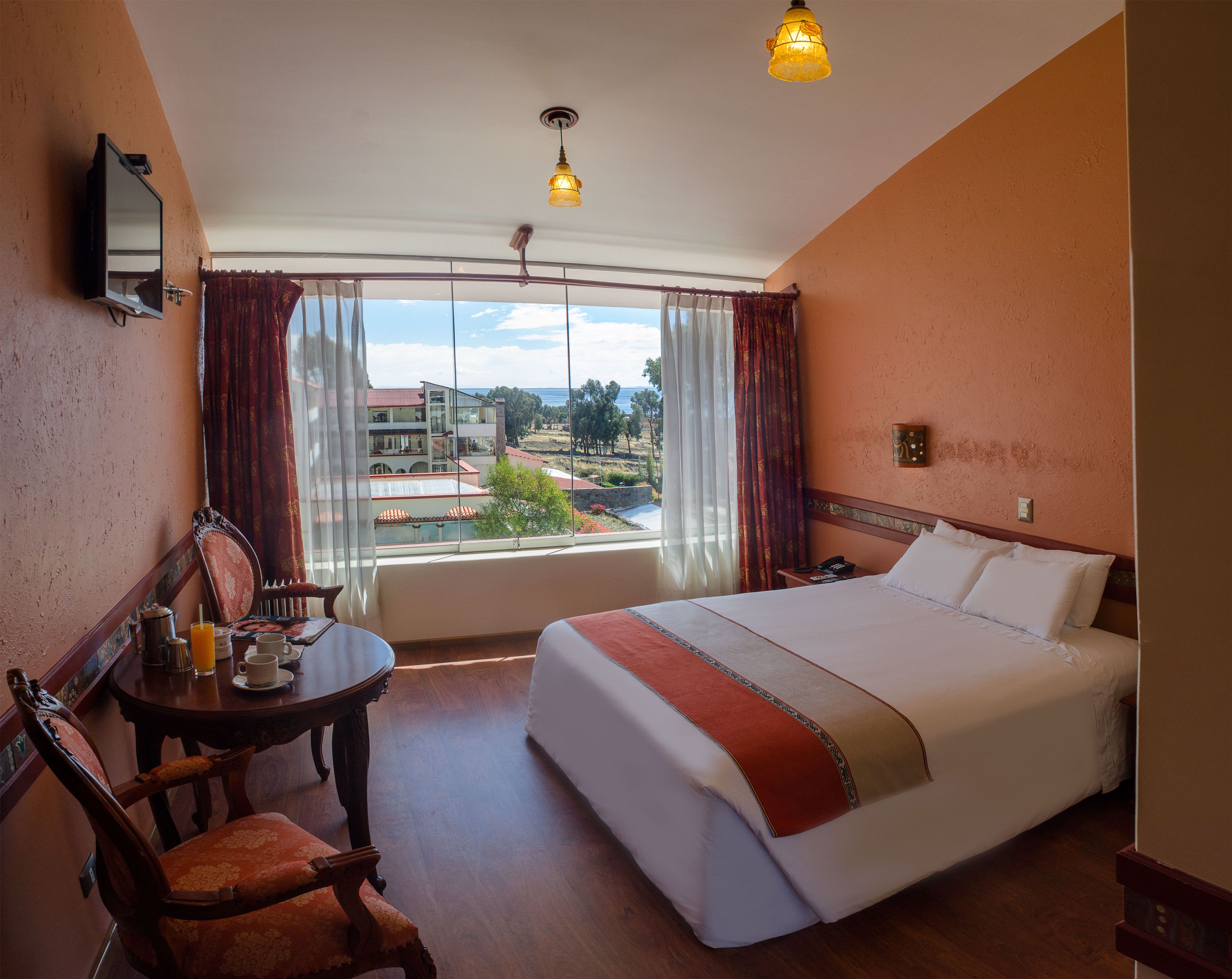 Habitación de hotel con cama, escritorio con silla, mesa con silla, ventana con cortinas y vistas a edificios.