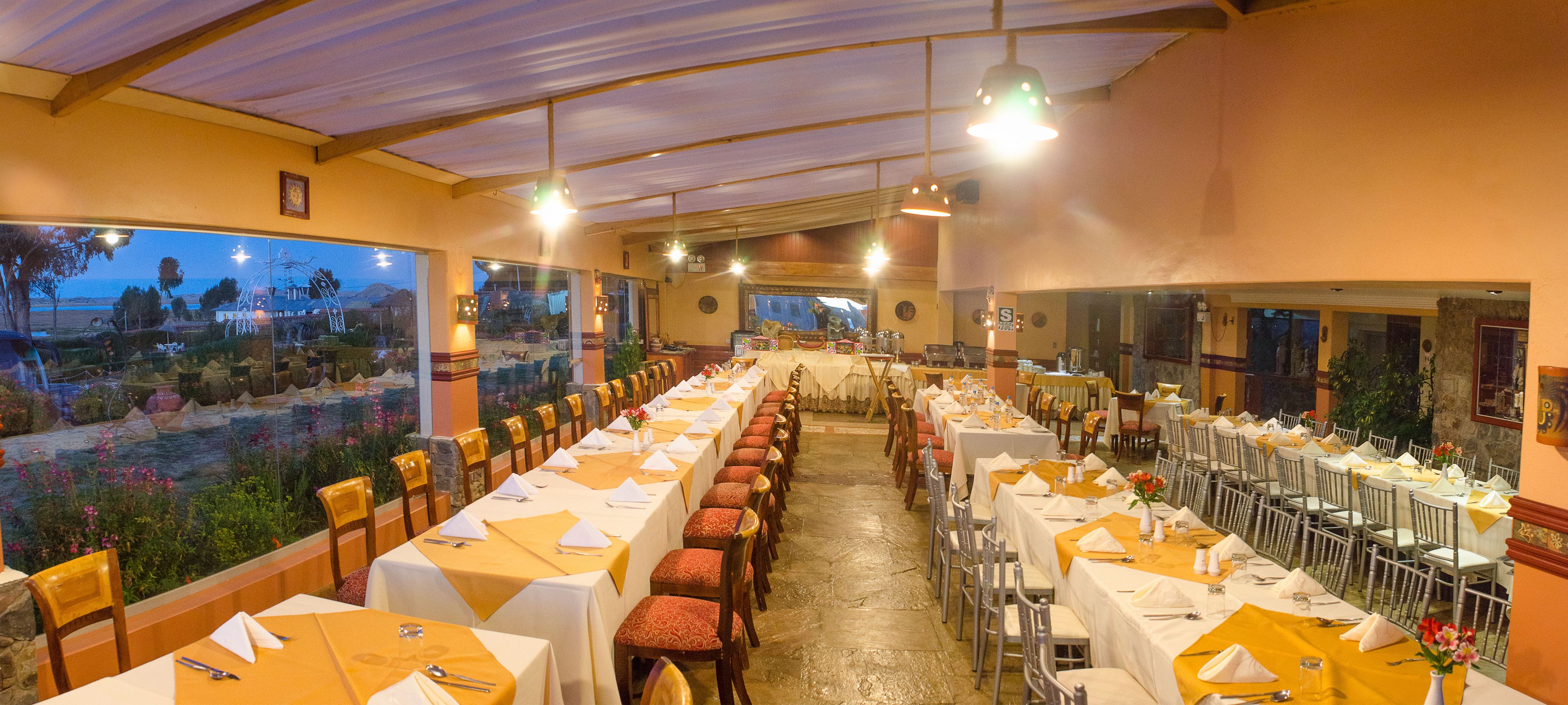 A long dining hall with tables set for a meal, large windows, and a view of the outdoors.