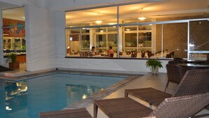 An indoor swimming pool with lounge chairs and a restaurant area with large windows. - Arituba Park Hotel (Natal)