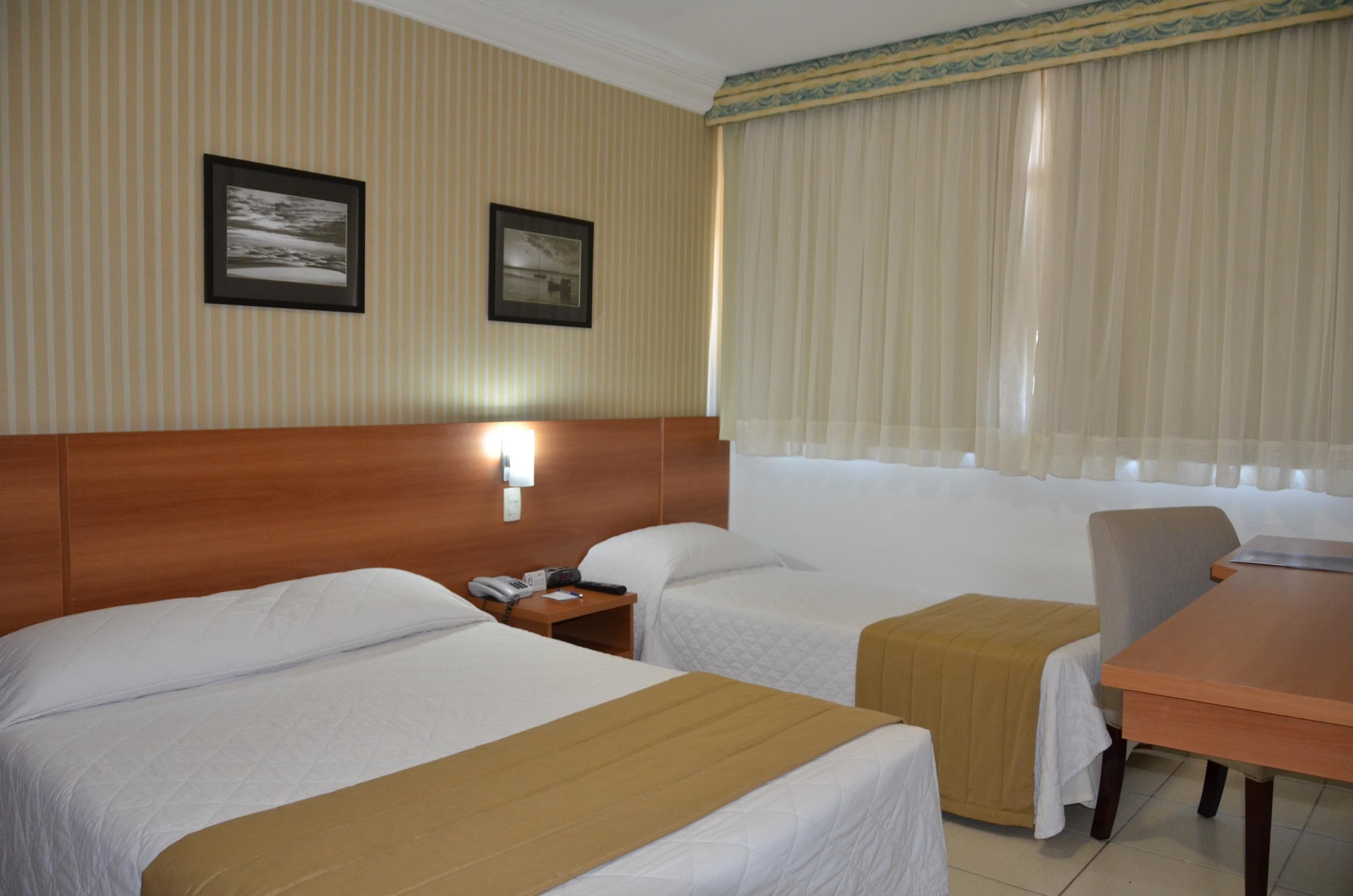 A hotel room with two beds, a wooden headboard, a small table, and a chair. There are two framed pictures on the wall.