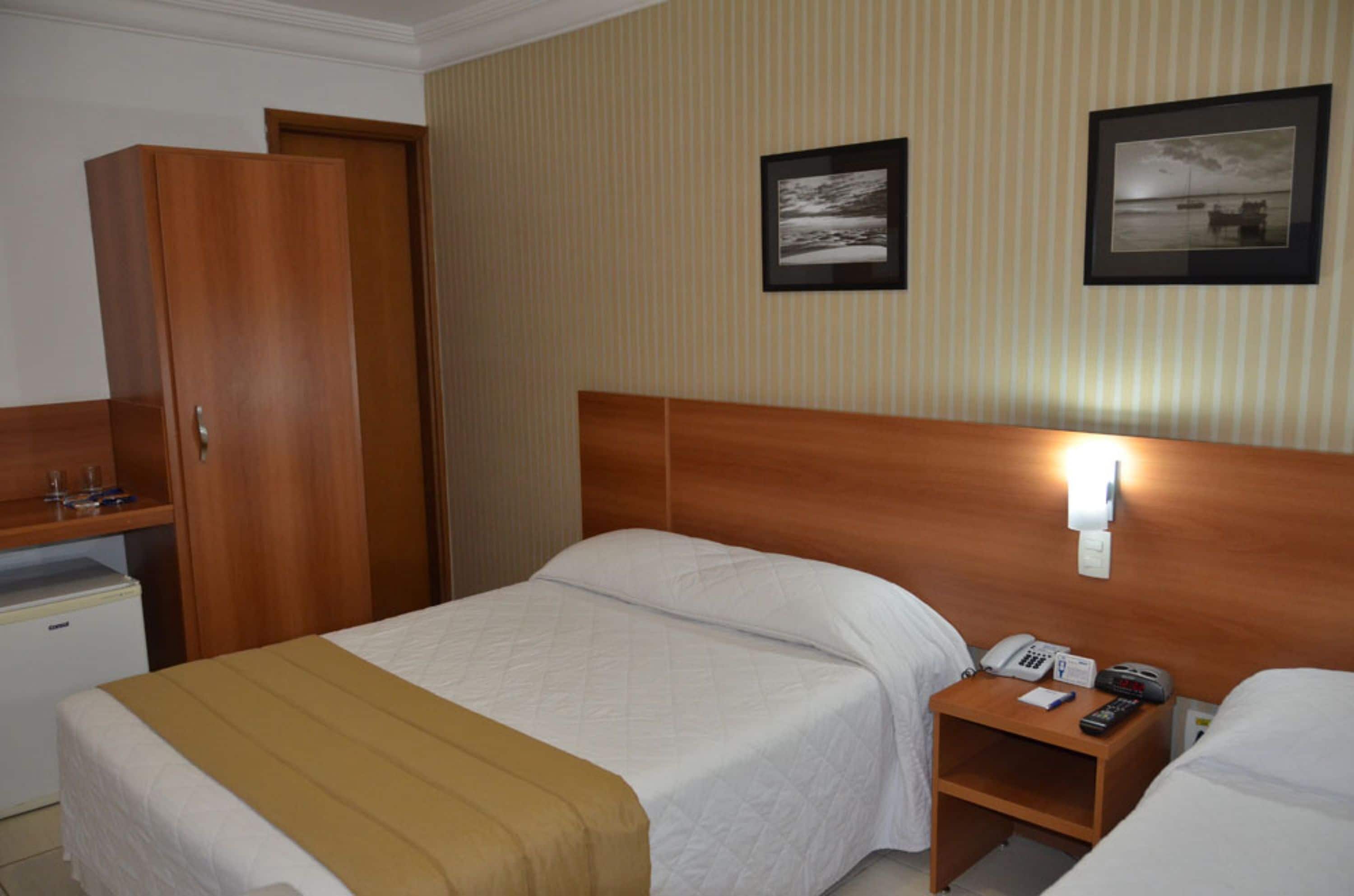 A hotel room with a bed, a bedside table with a phone and remote, a wardrobe, and a small desk with a refrigerator.
