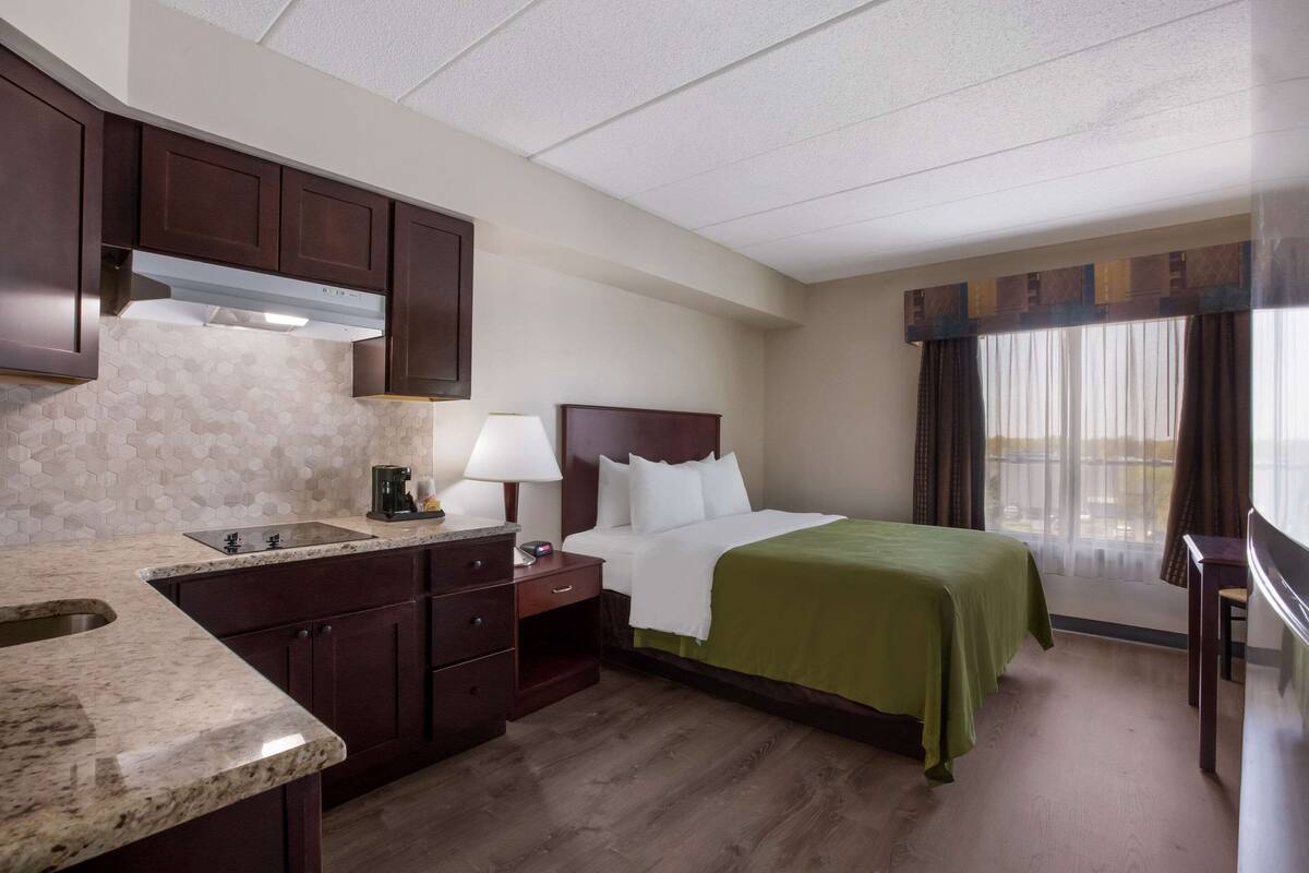 A hotel room with a kitchenette, a bed with a green bedspread, a nightstand, a lamp, and a view of the outdoors through the window.