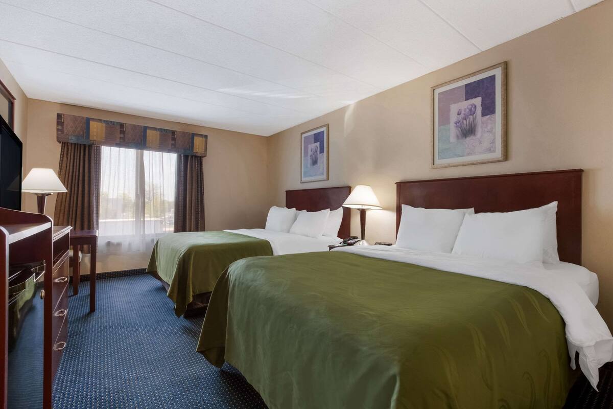 A hotel room with two beds, a desk, a lamp, and a window with curtains.