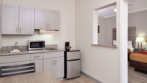 A compact kitchen with a microwave, coffee maker, and a small refrigerator. There is a dining table with chairs and a view of a bedroom through an open doorway. - Econo Lodge Inn & Suites Port Arthur near Sabine Pass (Port Arthur)