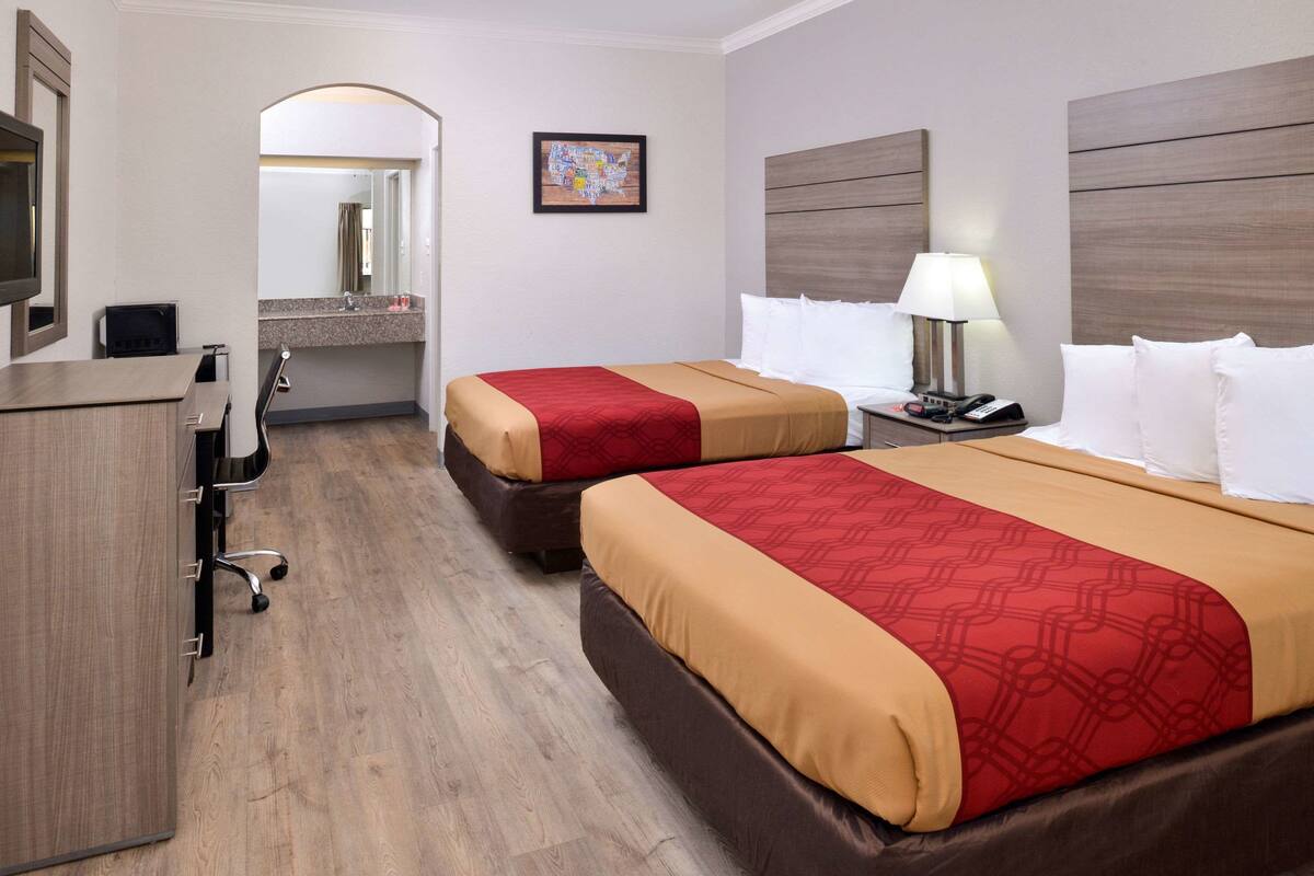 A hotel room with two beds, a desk, a chair, a TV, and a bathroom visible through an archway.