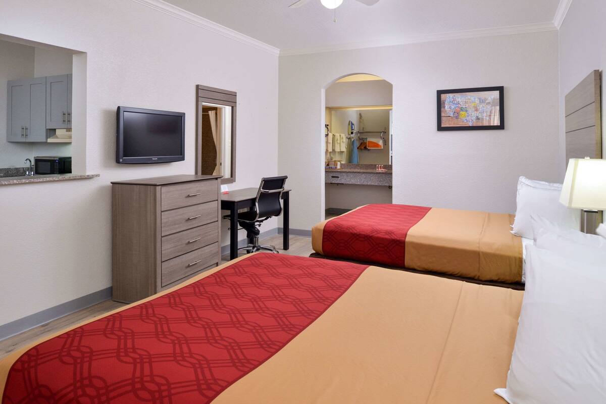 A hotel room with two beds, a desk, a chair, a TV, a microwave, and a bathroom visible through an archway.
