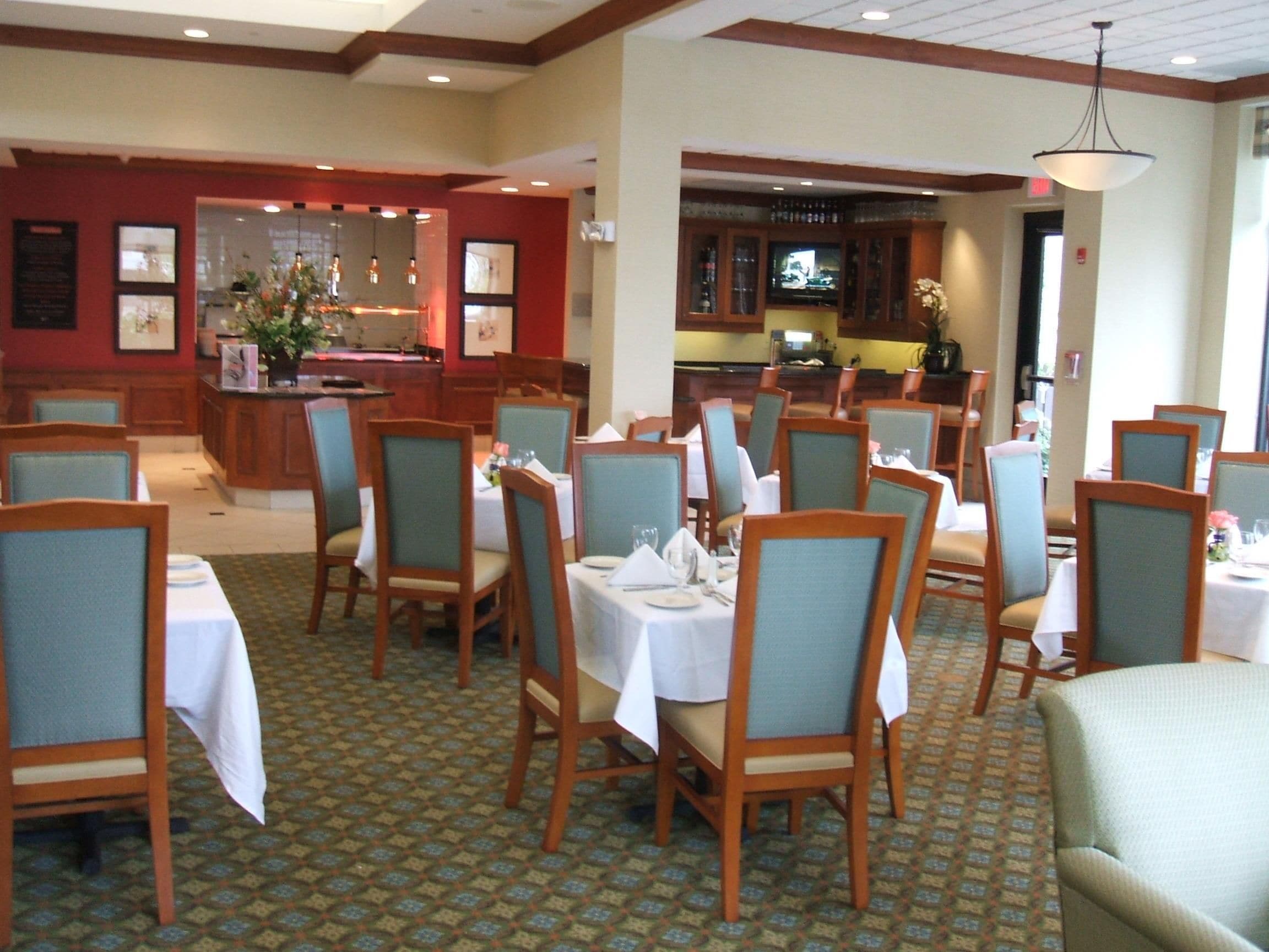 A hotel dining area with multiple tables and chairs, a bar area with a television, and a reception desk.