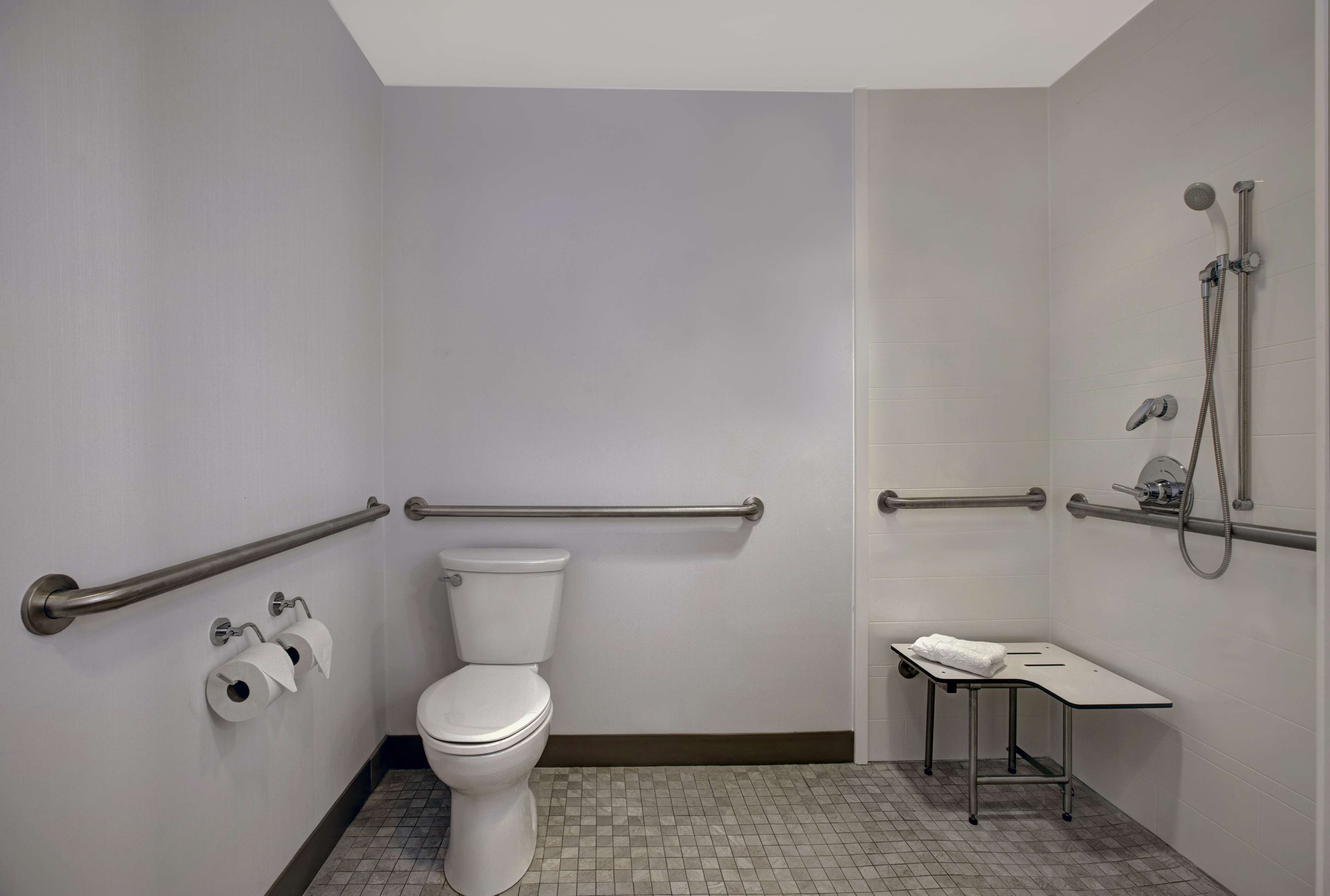 A bathroom with a toilet, handrails, a shower, and a small table with a towel.