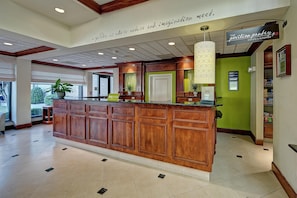 A hotel reception area with a wooden reception desk, a green accent wall, and a sign for 'The Pavilion Pantry.' - Hilton Garden Inn Memphis/Southaven, MS (Southaven)