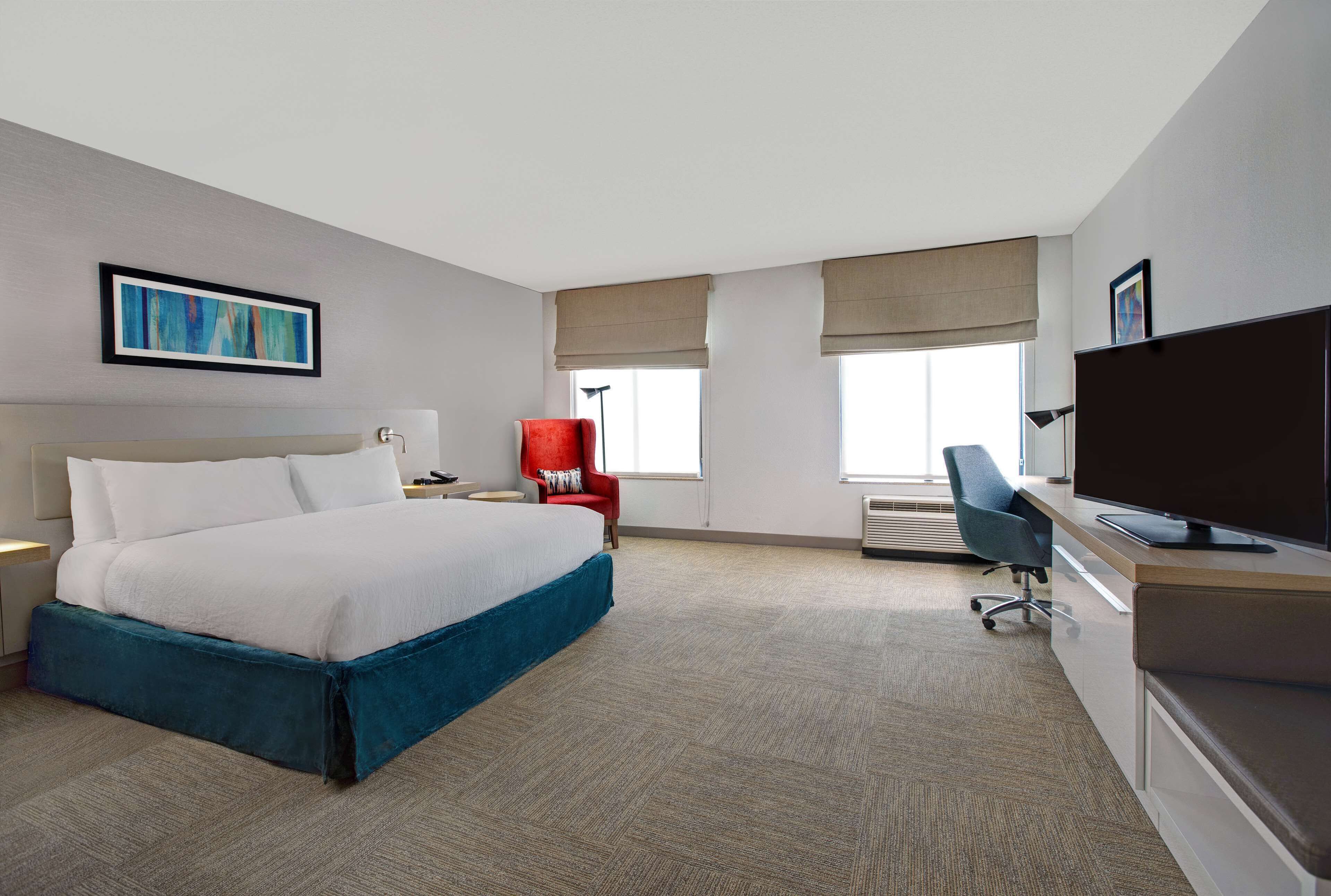 A hotel room with a large bed, a desk with a computer, a red chair, and a television.