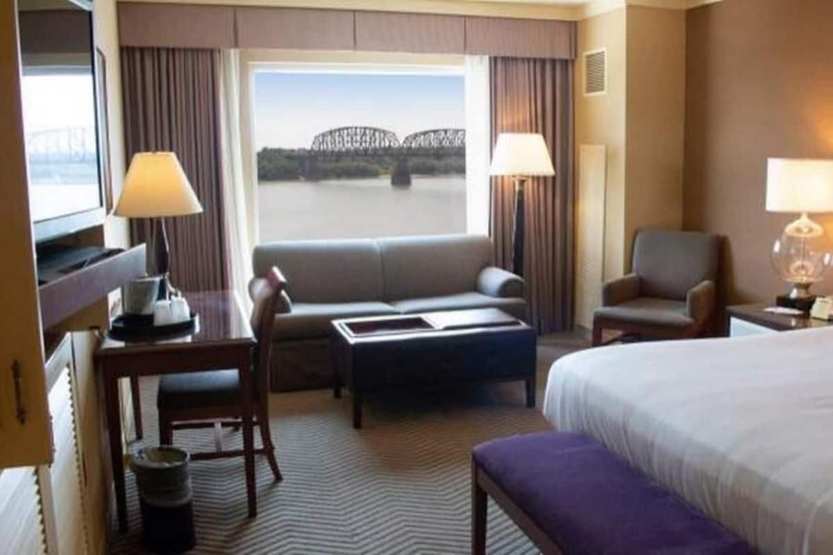 A hotel room with a bed, a sofa, a desk, a chair, a lamp, and a view of a bridge.
