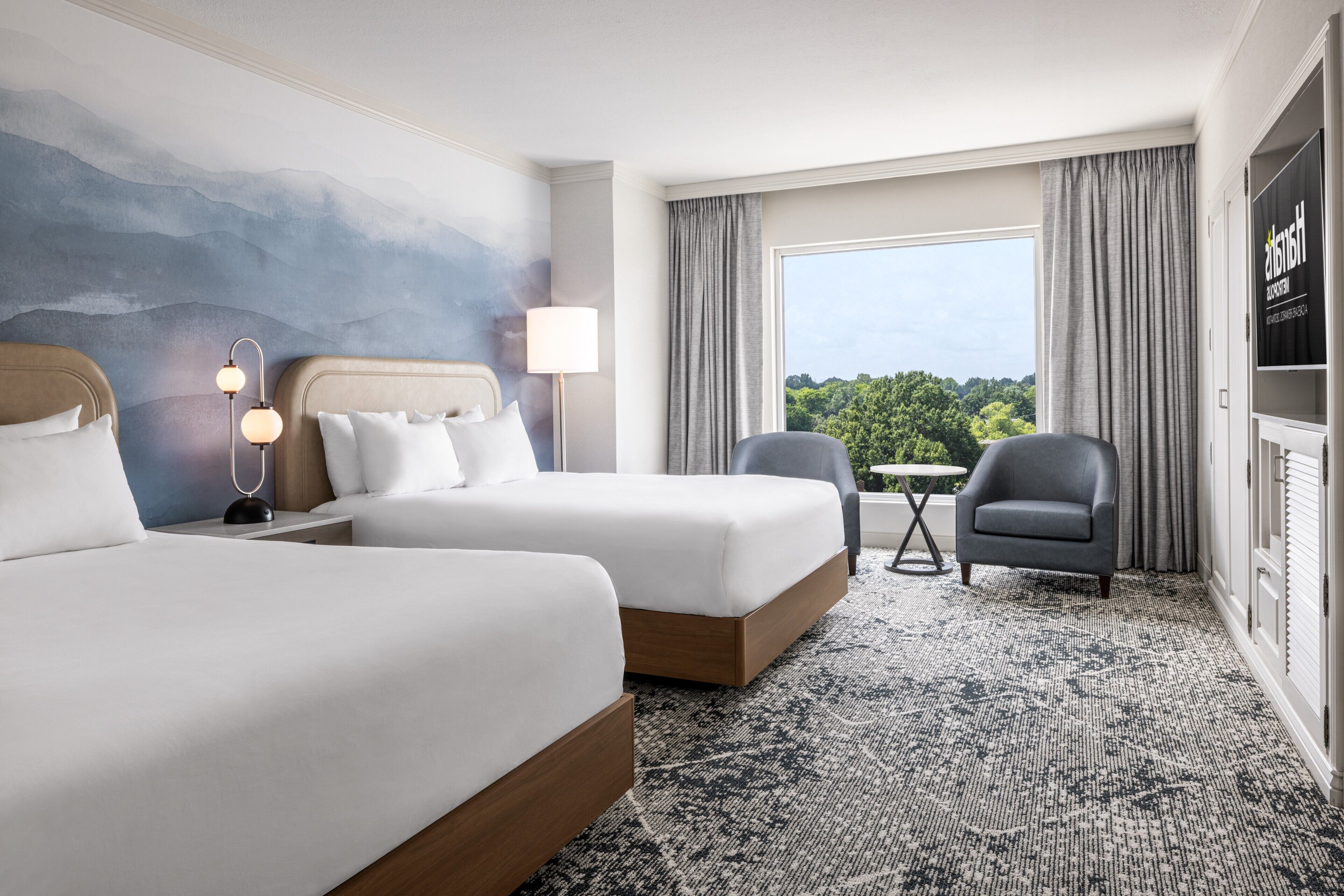 A hotel room with two beds, a desk, a chair, a lamp, and a view of greenery through the window.