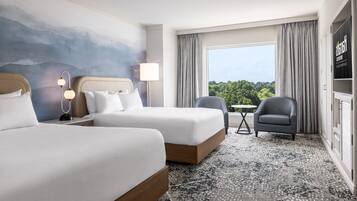 Une chambre d’hôtel avec deux lits, un bureau, une chaise, une lampe et une vue sur la verdure par la fenêtre.
