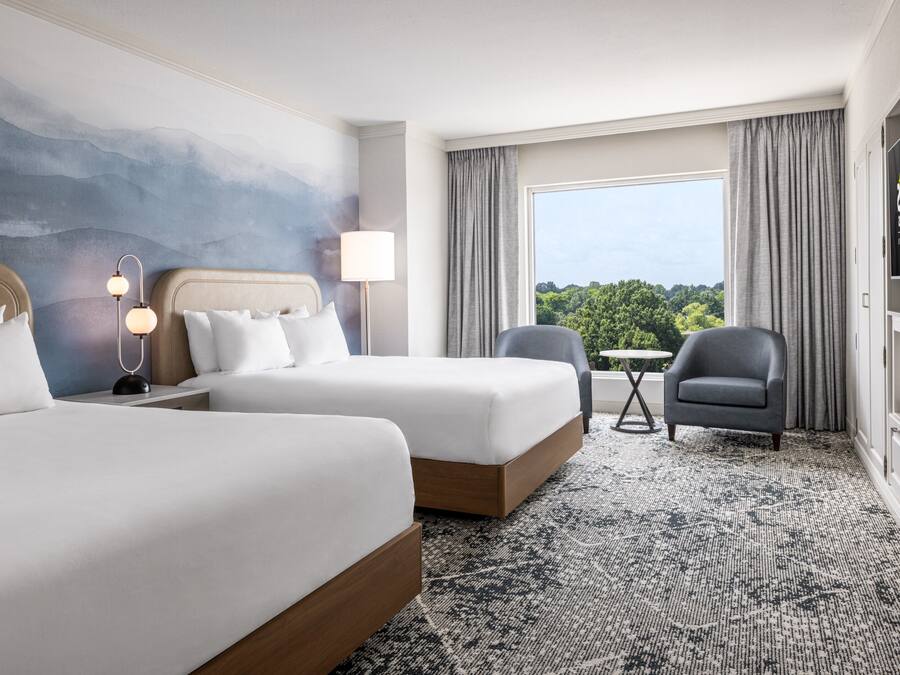 Une chambre d’hôtel avec deux lits, un bureau, une chaise, une lampe et une vue sur la verdure par la fenêtre.