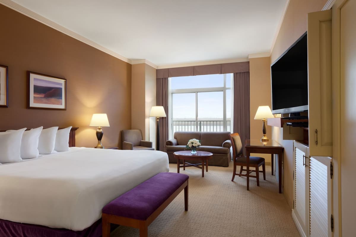 A hotel room with a large bed, a sofa, a small table, a bench, a TV, and a window with curtains.