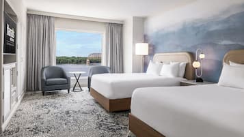 Quarto de hotel com duas camas, uma mesa, uma cadeira, um abajur e vista para uma ponte.