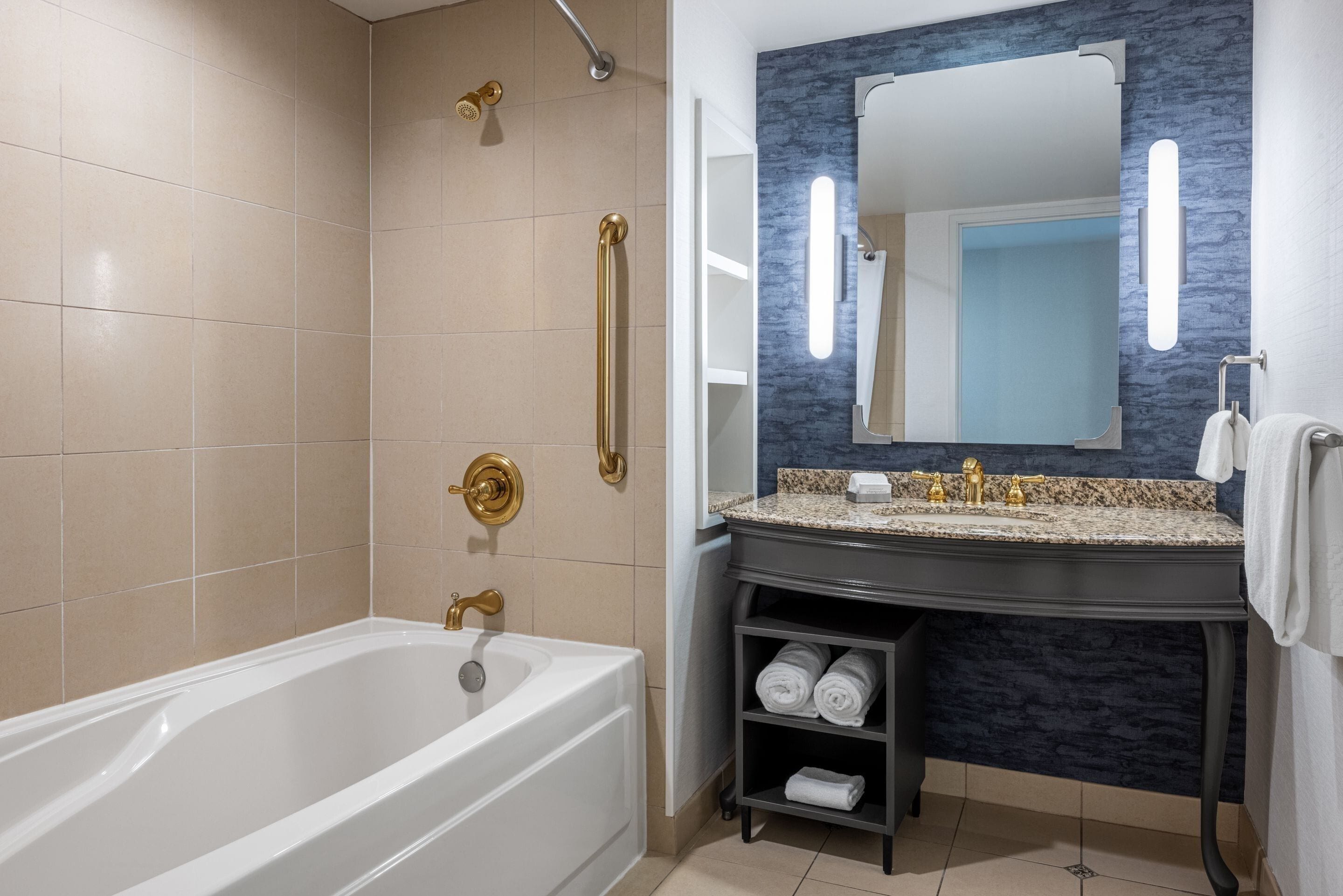 Een moderne badkamer met een bad, douche met gouden douchekop, wastafel met granieten blad en een spiegel met blauwe accentmuur.