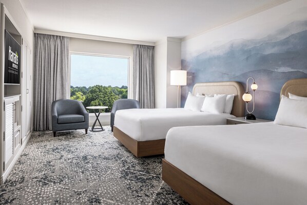 A hotel room with two beds, a desk, a chair, a view of the outdoors, and a painting on the wall.