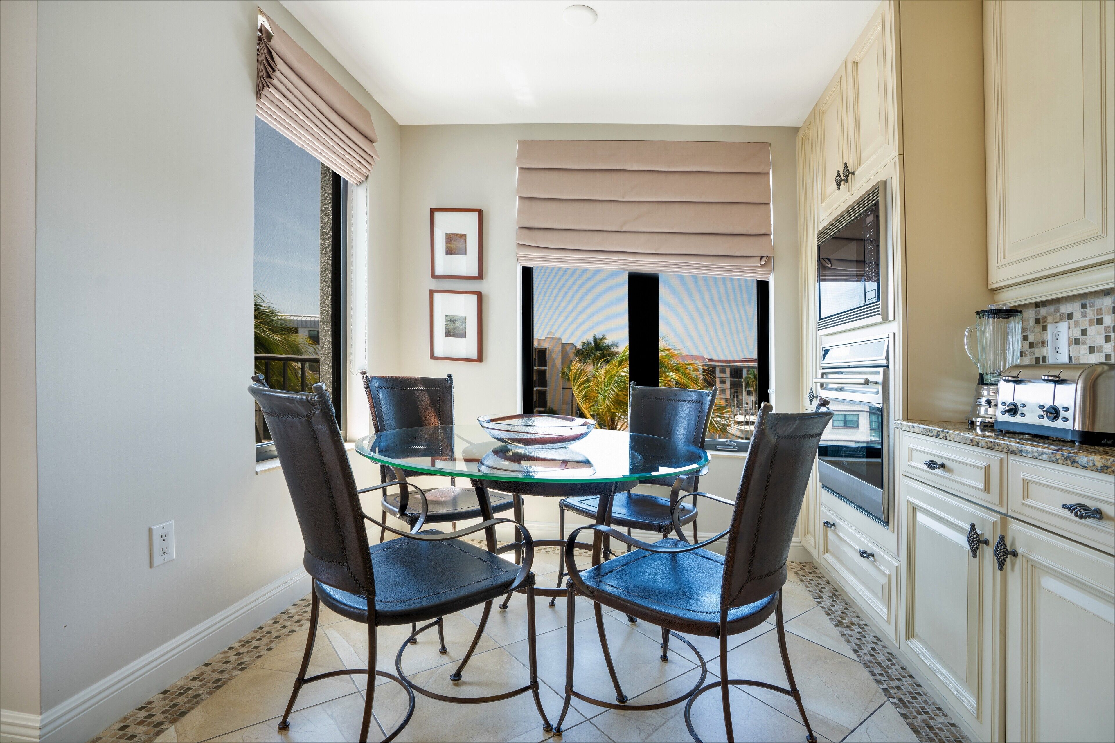 A dining area with a glass table and four chairs, a kitchen with white cabinets and appliances, and a view of the outdoors through large windows.