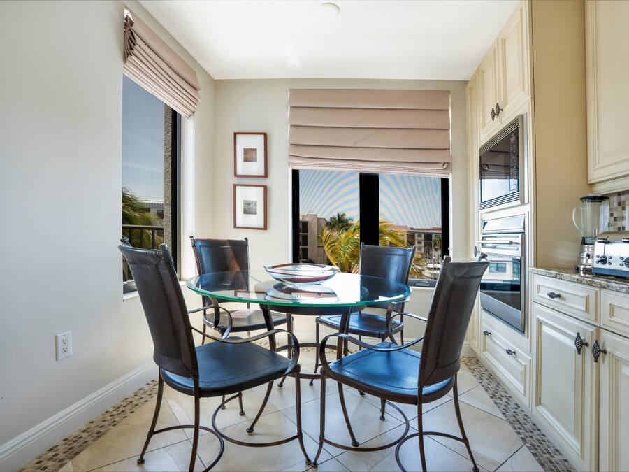A dining area with a glass table and four chairs, a kitchen with white cabinets and appliances, and a view of the outdoors through large windows.