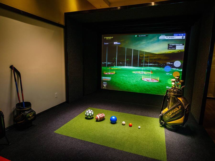 A room with a large screen displaying a golf video game, a golf bag, and various sports balls on a green mat.