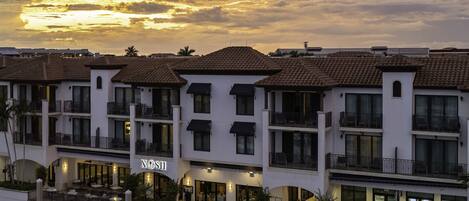 Un hotel frente al mar con varios balcones, un puerto deportivo y vistas al atardecer.