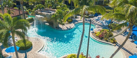 Piscina de resort con cascada, rodeada de palmeras y sillones de descanso.