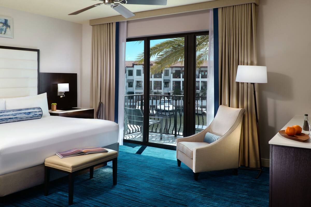 A hotel room with a large bed, a desk, a chair, and a balcony with a view of buildings and palm trees.