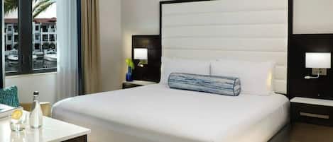 A hotel room with a large bed, two bedside tables, a nightstand, a window with a view of boats, and a painting on the wall.