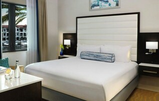 A hotel room with a large bed, two bedside tables, a nightstand, a window with a view of boats, and a painting on the wall.