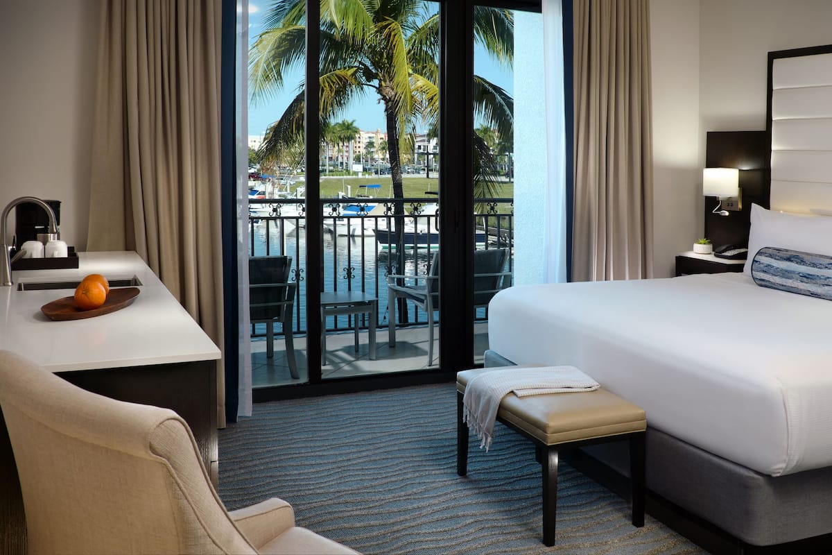 A hotel room with a bed, a desk, a chair, a small table with an orange, and a view of a pool and palm trees.