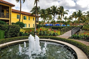 A fountain surrounded by a landscaped garden, a bridge, and a yellow building with a balcony.